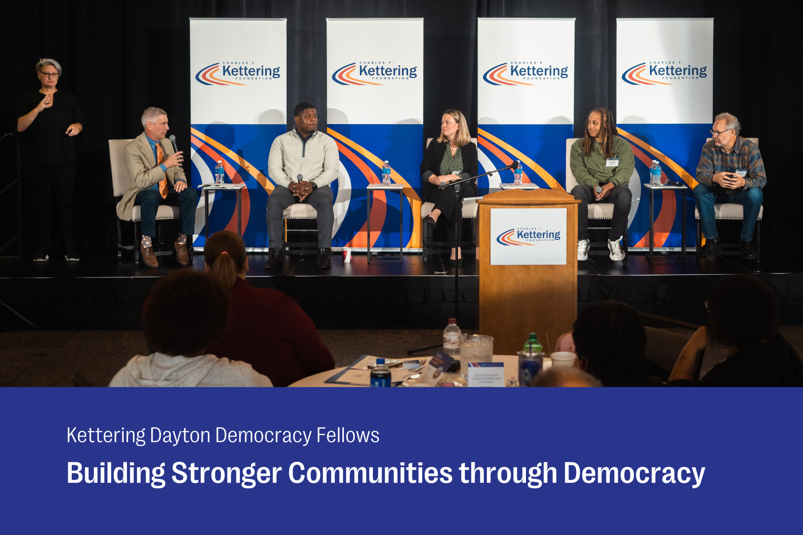 A panel of five speakers, including three men and two women, sits on stage at the Kettering Foundation's Dayton Democracy Fellows event. The backdrop displays the Kettering Foundation logo, with an American Sign Language interpreter standing to the left. Attendees are seated in the audience, engaging with the event, and the room's atmosphere is professional. The banner below the stage reads 'Building Stronger Communities through Democracy.